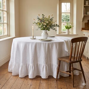Puede incluir: Una mantel redondo de lino blanco con borde con volantes cubre una mesa de madera. Un jarrón con flores está en el centro, con una vela encendida y una silla de madera cerca. La habitación tiene un ambiente acogedor.