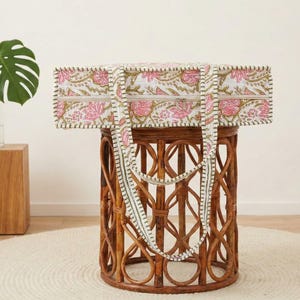 May include: A rectangular tote bag with a floral pattern in pink and white, with green accents. The bag has a matching strap and is displayed on a brown wicker table. A green plant in a glass vase sits on a wooden block.