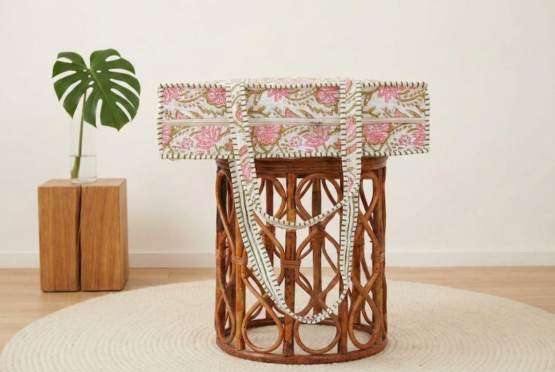 May include: A rectangular tote bag with a floral pattern in pink and white, with green accents. The bag has a matching strap and is displayed on a brown wicker table. A green plant in a glass vase sits on a wooden block.