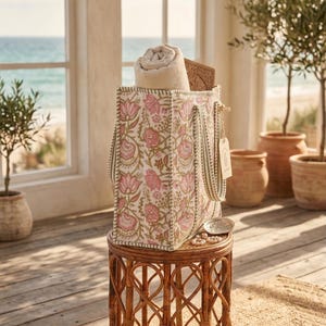 May include: A floral print tote bag with pink and green accents sits on a woven side table. The bag contains a rolled-up beige towel and a wooden box. The scene is set near the ocean, with olive trees in terracotta pots.