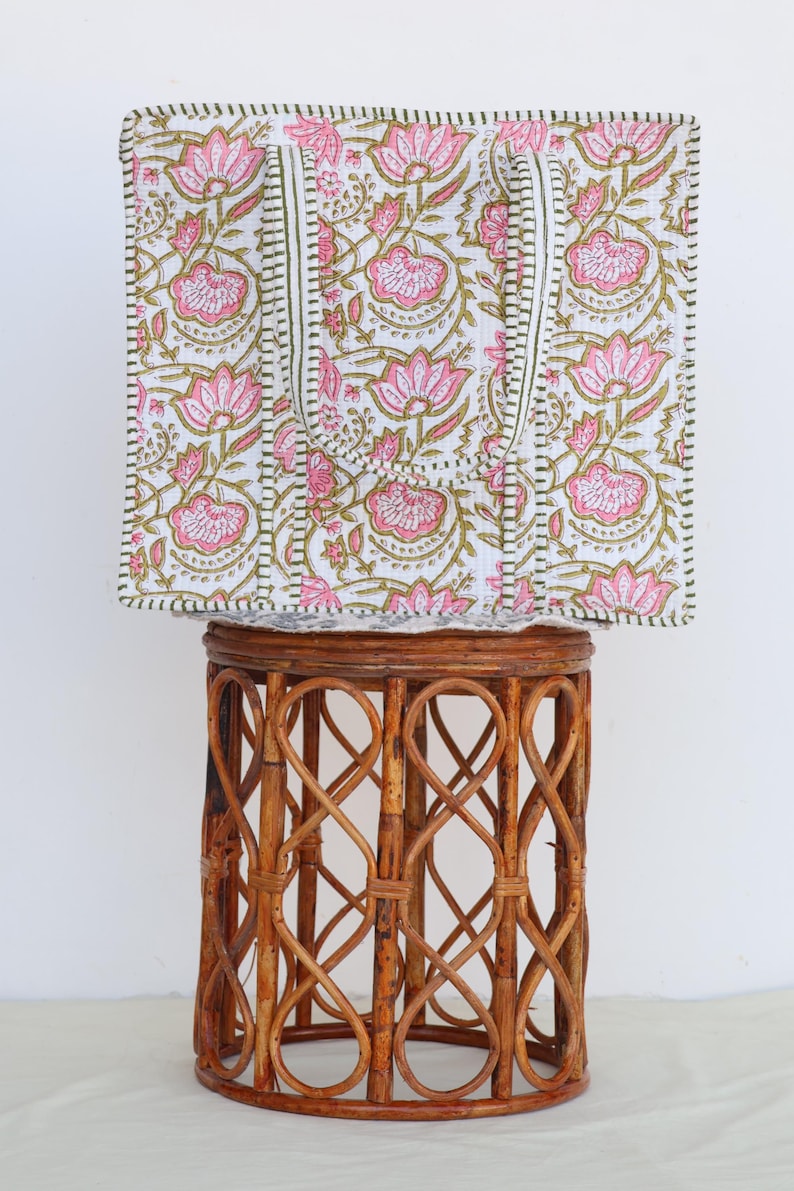 May include: A square tote bag with a floral pattern in pink and olive green on a white background. The bag has two white handles and is sitting on a brown wicker stool. The bag's edges are trimmed with a dark green border.