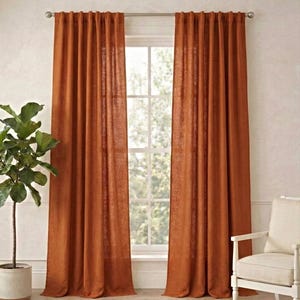 May include: Two rust-coloured linen curtains hang on a silver rod in front of a window. A potted plant and a white chair with a cream-coloured cushion are on either side of the window. A woven rug covers the floor.