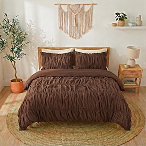 May include: A brown bedspread and pillow set with a ruched design. The bedding is on a wooden bed frame, with a woven rug beneath. A macrame wall hanging and a small side table with a lamp complete the bedroom decor.