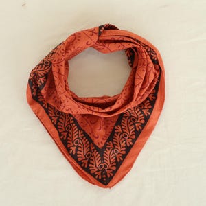 Könnte beinhalten: Ein orangefarbenes Bandana mit einem schwarzen Blumenmuster. Das Bandana ist gefaltet und gebunden und zeigt das komplizierte Muster entlang der Kanten. Der Stoff wirkt weich und leicht und eignet sich für verschiedene Zwecke.