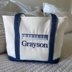 Pode incluir: Bolsa tote creme com detalhes em azul marinho, incluindo as alças e a base. A bolsa apresenta um design bordado de teclado de piano acima do nome "Grayson" em azul marinho. A bolsa está sobre uma colcha branca acolchoada.