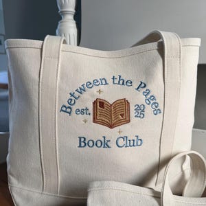 Pode incluir: Uma bolsa tote de lona cor de creme com texto bordado em azul que diz Between the Pages Book Club, est. 2025 e um &iacute;cone central de um livro aberto bordado em marrom rodeado por pequenas estrelas douradas.
