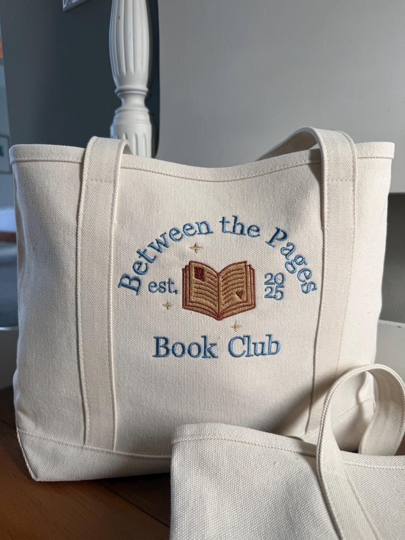 Pode incluir: Uma bolsa tote de lona cor de creme com texto bordado em azul que diz Between the Pages Book Club, est. 2025 e um &iacute;cone central de um livro aberto bordado em marrom rodeado por pequenas estrelas douradas.
