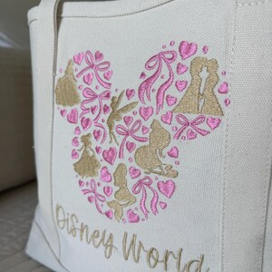 May include: Off-white canvas tote bag with a Mickey Mouse head design filled with pink hearts, bows, and gold silhouettes of Disney characters. The words "Disney World" are embroidered in gold.