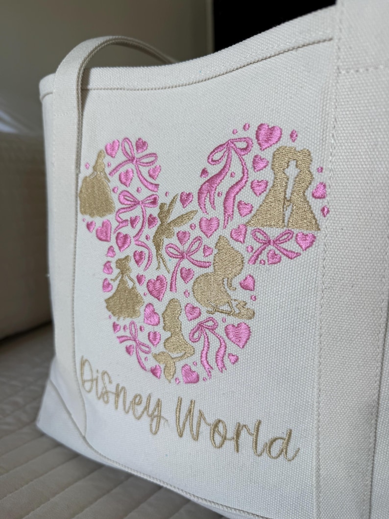 May include: Off-white canvas tote bag with a Mickey Mouse head design filled with pink hearts, bows, and gold silhouettes of Disney characters. The words "Disney World" are embroidered in gold.