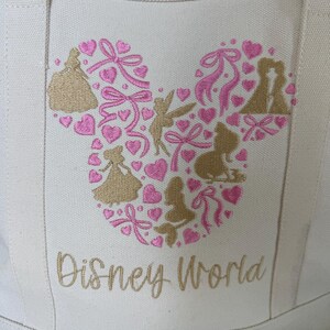 May include: Off-white canvas tote bag with a Mickey Mouse head design made of pink hearts, bows, and gold silhouettes of Disney characters. The words "Disney World" are embroidered in gold script below the design.