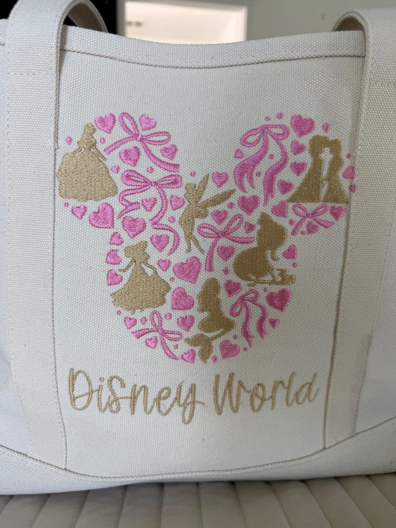 May include: Off-white canvas tote bag with a Mickey Mouse head design made of pink hearts, bows, and gold silhouettes of Disney characters. The words "Disney World" are embroidered in gold script below the design.