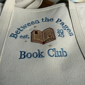 Pode incluir: Uma bolsa tote de lona na cor off-white com texto bordado em azul que diz Between the Pages Book Club. O design inclui uma ilustra&ccedil;&atilde;o de um livro aberto com um pequeno cora&ccedil;&atilde;o vermelho, a abrevia&ccedil;&atilde;o est. e o ano 2025 em fio azul.