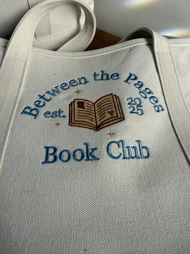 Pode incluir: Uma bolsa tote de lona na cor off-white com texto bordado em azul que diz Between the Pages Book Club. O design inclui uma ilustra&ccedil;&atilde;o de um livro aberto com um pequeno cora&ccedil;&atilde;o vermelho, a abrevia&ccedil;&atilde;o est. e o ano 2025 em fio azul.