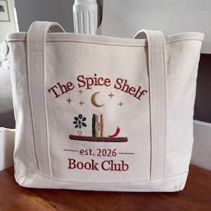 Pode incluir: Uma sacola de lona na cor off-white com o texto bordado The Spice Shelf Book Club est. 2026. O design inclui uma prateleira marrom com uma planta em vaso, dois livros, uma pimenta vermelha, uma lua crescente dourada e pequenas estrelas.