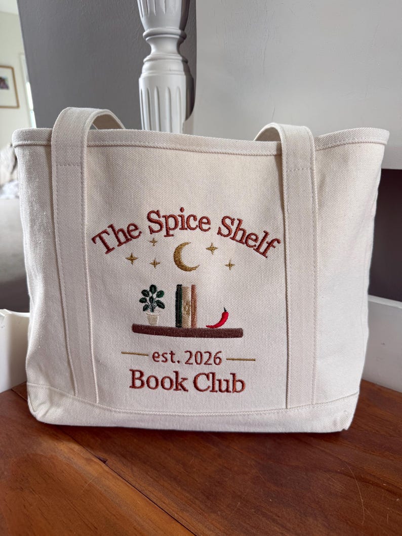 Pode incluir: Uma sacola de lona na cor off-white com o texto bordado The Spice Shelf Book Club est. 2026. O design inclui uma prateleira marrom com uma planta em vaso, dois livros, uma pimenta vermelha, uma lua crescente dourada e pequenas estrelas.