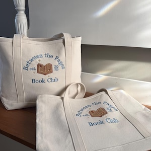 Pode incluir: Duas sacolas de lona na cor off-white com texto bordado em azul que diz Between the Pages est. 2025 Book Club, acima de um &iacute;cone de livro aberto bordado em marrom com pequenos detalhes de estrelas douradas na frente.
