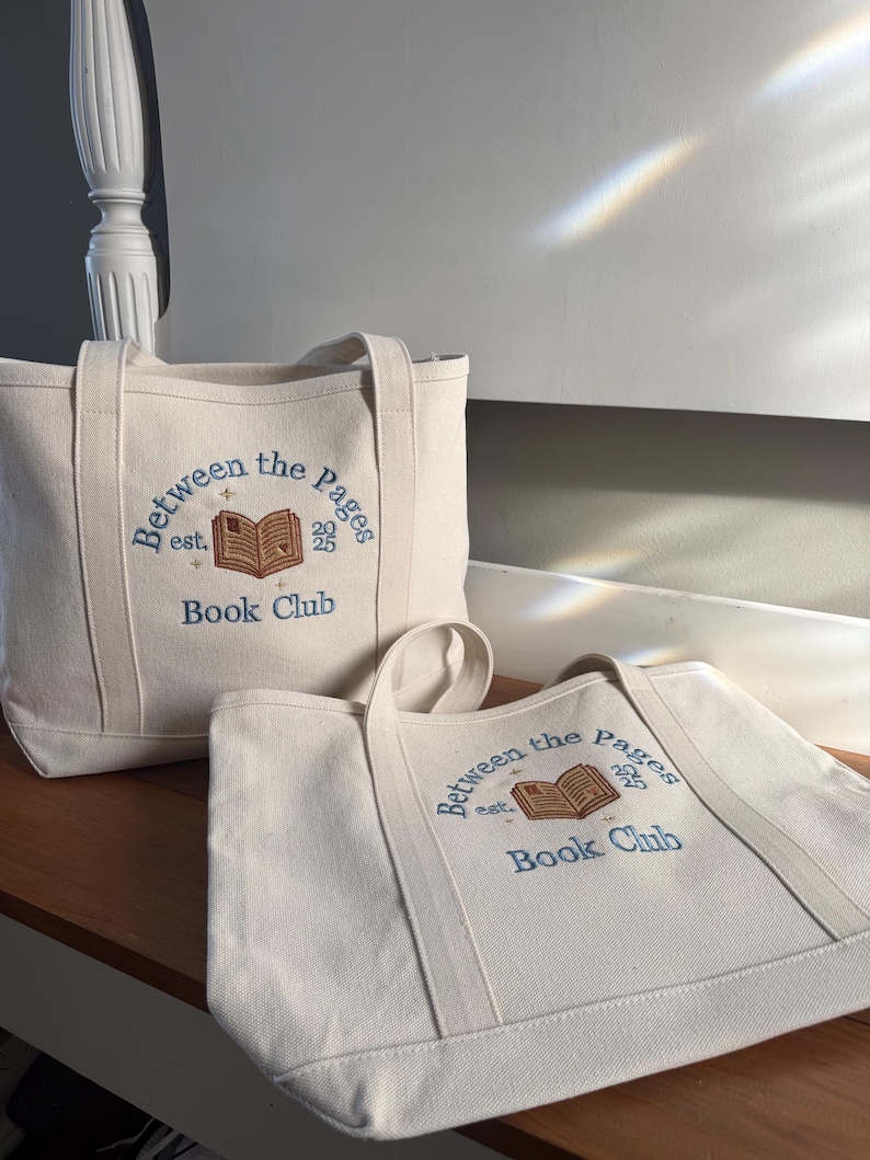 Pode incluir: Duas sacolas de lona na cor off-white com texto bordado em azul que diz Between the Pages est. 2025 Book Club, acima de um &iacute;cone de livro aberto bordado em marrom com pequenos detalhes de estrelas douradas na frente.