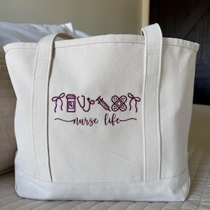 May include: Off-white canvas tote bag with two handles. The bag features an embroidered design in a deep purple color, including medical symbols and the words "nurse life". The bag is sitting on a bed.