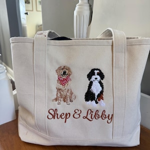 Pode incluir: Uma bolsa tote de lona na cor creme com ilustrações bordadas de dois cães. Um cão é um retriever dourado com uma bandana vermelha, e o outro é um cão preto, branco e castanho. O texto Shep & Libby está bordado por baixo.