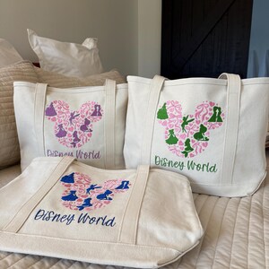 May include: Three off-white canvas tote bags with embroidered Disney World designs. Each bag features a Mickey Mouse head shape filled with silhouettes and floral patterns in pink, blue, and green. The words "Disney World" are embroidered in blue or green.