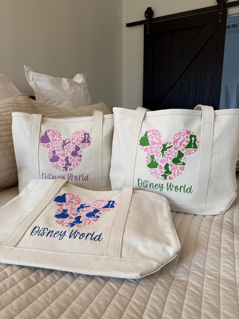May include: Three off-white canvas tote bags with embroidered Disney World designs. Each bag features a Mickey Mouse head shape filled with silhouettes and floral patterns in pink, blue, and green. The words "Disney World" are embroidered in blue or green.