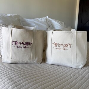 May include: Two off-white canvas tote bags with matching handles. Each bag has a purple embroidered design of medical tools and the words "nurse life". The bags are sitting on a bed with a white comforter.
