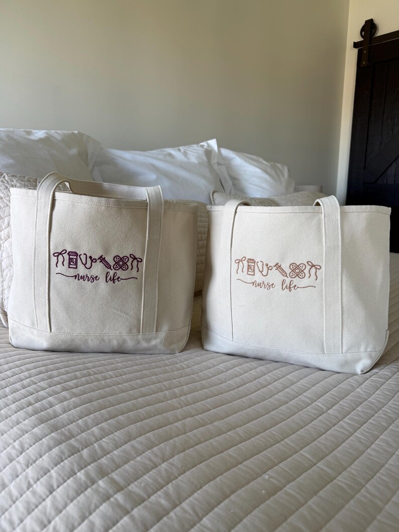 May include: Two off-white canvas tote bags with matching handles. Each bag has a purple embroidered design of medical tools and the words "nurse life". The bags are sitting on a bed with a white comforter.