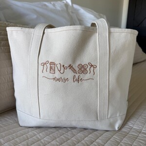 May include: Off-white canvas tote bag with brown embroidered medical-themed icons and the words "nurse life." The bag has two handles and is sitting on a bed. The embroidery includes a pill bottle, stethoscope, syringe, bandage, and a heart.