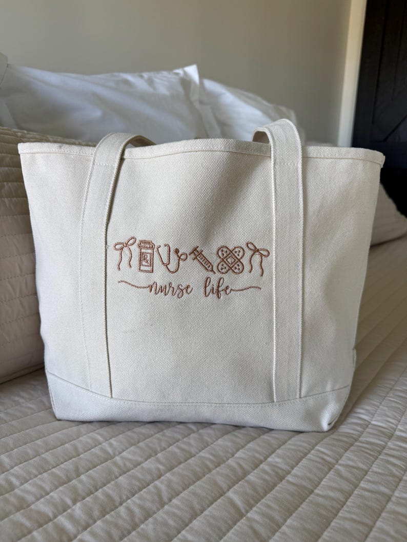 May include: Off-white canvas tote bag with brown embroidered medical-themed icons and the words "nurse life." The bag has two handles and is sitting on a bed. The embroidery includes a pill bottle, stethoscope, syringe, bandage, and a heart.