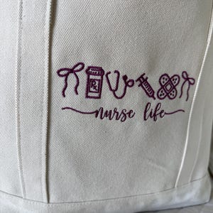 May include: Off-white canvas tote bag with embroidered purple medical symbols and the words "nurse life." The design includes a syringe, stethoscope, pill bottle, and bandage. The bag has a simple, functional design.