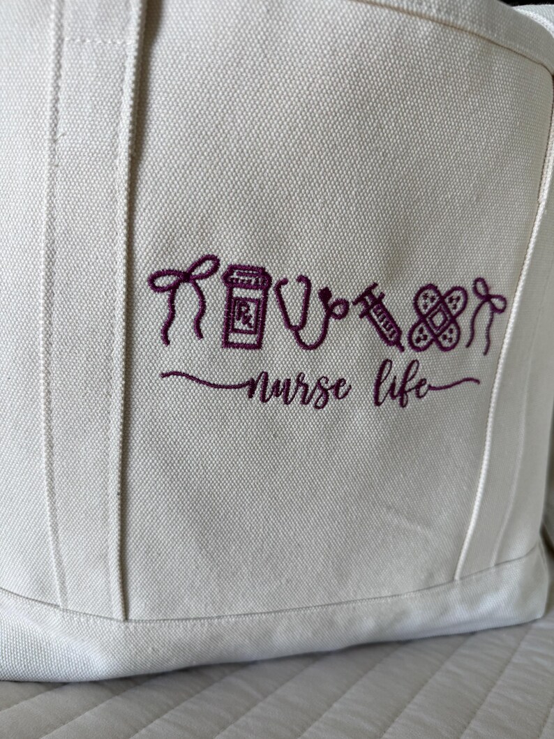 May include: Off-white canvas tote bag with embroidered purple medical symbols and the words "nurse life." The design includes a syringe, stethoscope, pill bottle, and bandage. The bag has a simple, functional design.