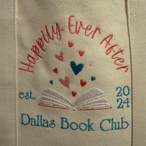 Pode incluir: Uma bolsa de lona cor de creme com bordado que diz Happily Ever After, est. 2024 e Dallas Book Club. Um gr&aacute;fico de um livro aberto est&aacute; centralizado com pequenos cora&ccedil;&otilde;es vermelhos, rosas e azul-petr&oacute;leo sobre as p&aacute;ginas.