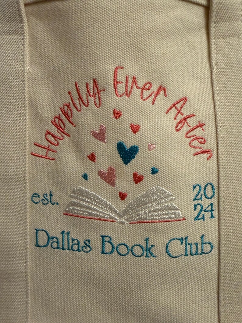 Pode incluir: Uma bolsa de lona cor de creme com bordado que diz Happily Ever After, est. 2024 e Dallas Book Club. Um gr&aacute;fico de um livro aberto est&aacute; centralizado com pequenos cora&ccedil;&otilde;es vermelhos, rosas e azul-petr&oacute;leo sobre as p&aacute;ginas.