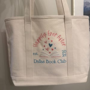 Pode incluir: Uma sacola de lona cor de creme com texto bordado que diz "Happily Ever After" e "Dallas Book Club" com "est. 2024". O design inclui um gr&aacute;fico de um livro aberto com pequenos cora&ccedil;&otilde;es vermelhos, rosas e azuis flutuando.