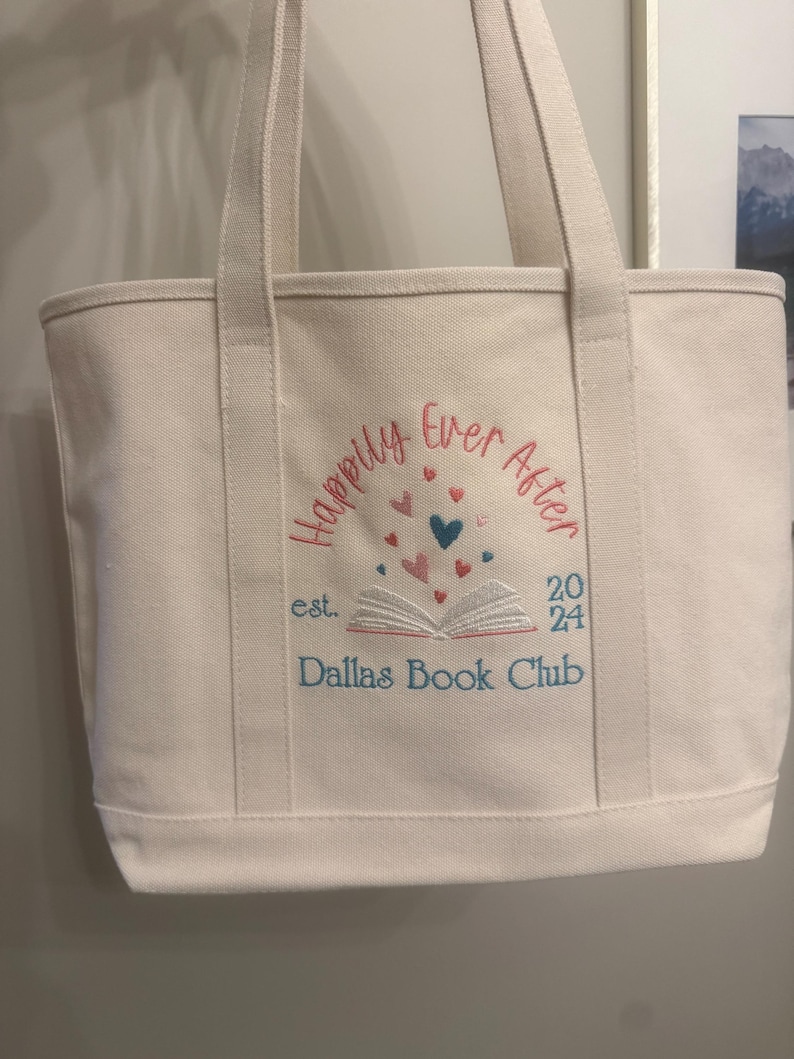 Pode incluir: Uma sacola de lona cor de creme com texto bordado que diz "Happily Ever After" e "Dallas Book Club" com "est. 2024". O design inclui um gr&aacute;fico de um livro aberto com pequenos cora&ccedil;&otilde;es vermelhos, rosas e azuis flutuando.