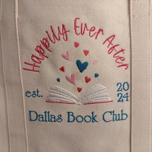 Pode incluir: Uma sacola de lona bege com texto bordado que diz Happily Ever After, est. 2024 e Dallas Book Club. Um gr&aacute;fico de um livro aberto est&aacute; centralizado com pequenos cora&ccedil;&otilde;es vermelhos, rosas e azuis flutuando acima das p&aacute;ginas.