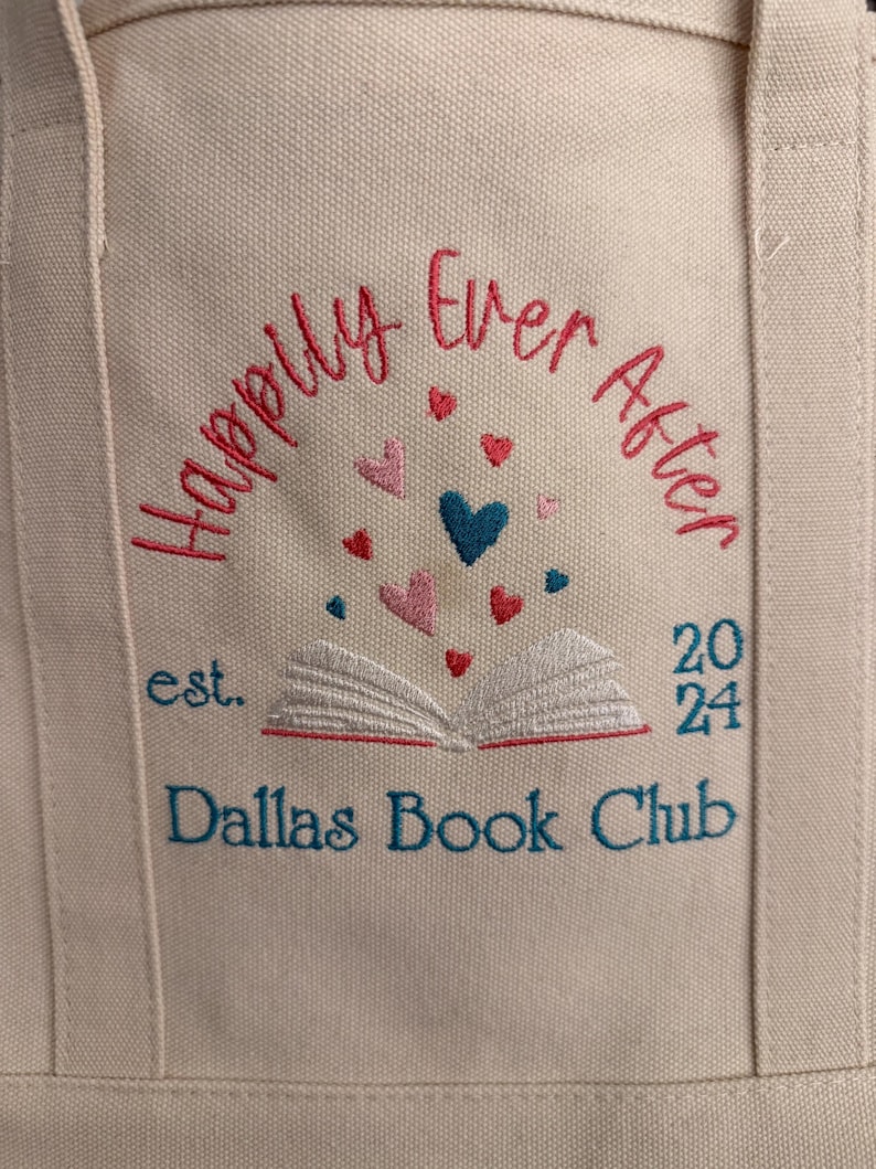 Pode incluir: Uma sacola de lona bege com texto bordado que diz Happily Ever After, est. 2024 e Dallas Book Club. Um gr&aacute;fico de um livro aberto est&aacute; centralizado com pequenos cora&ccedil;&otilde;es vermelhos, rosas e azuis flutuando acima das p&aacute;ginas.
