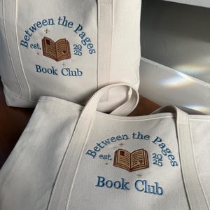 Pode incluir: Duas sacolas de lona na cor off-white com bordado azul que diz Between the Pages Book Club est. 2025. O design inclui um &iacute;cone de livro aberto marrom com pequenas estrelas douradas e um detalhe de cora&ccedil;&atilde;o na p&aacute;gina.