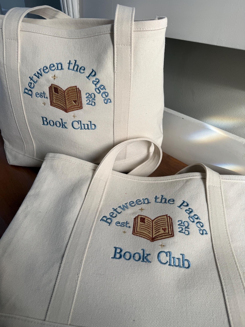 Pode incluir: Duas sacolas de lona na cor off-white com bordado azul que diz Between the Pages Book Club est. 2025. O design inclui um &iacute;cone de livro aberto marrom com pequenas estrelas douradas e um detalhe de cora&ccedil;&atilde;o na p&aacute;gina.