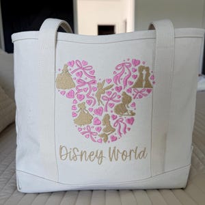 May include: Off-white canvas tote bag with a gold and pink Mickey Mouse head design. The design is filled with silhouettes of Disney characters and hearts. The words "Disney World" are embroidered in gold.
