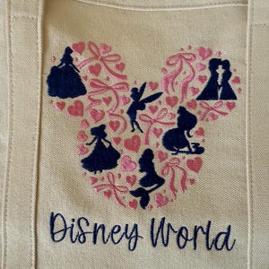 May include: Beige canvas tote bag featuring a pink and navy blue Mickey Mouse head design filled with silhouettes of Disney characters and pink hearts. The words "Disney World" are embroidered in navy blue.