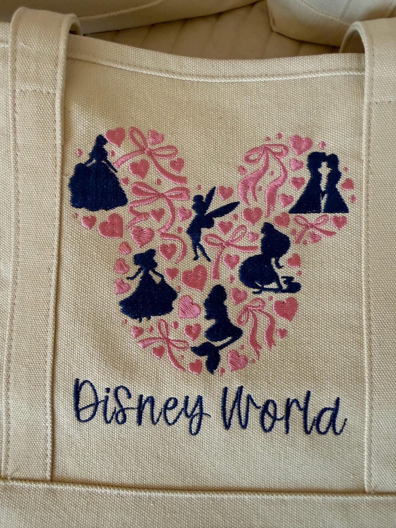 May include: Beige canvas tote bag featuring a pink and navy blue Mickey Mouse head design filled with silhouettes of Disney characters and pink hearts. The words "Disney World" are embroidered in navy blue.
