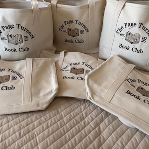 Pode incluir: Um conjunto de cinco sacolas de lona na cor branco off-white com bordado preto que diz The Page Turners est. 2025 Book Club, acompanhado por um gr&aacute;fico central de um livro aberto com um pequeno detalhe de cora&ccedil;&atilde;o e estrelas.