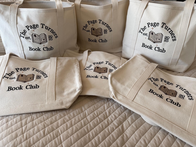 Pode incluir: Um conjunto de cinco sacolas de lona na cor branco off-white com bordado preto que diz The Page Turners est. 2025 Book Club, acompanhado por um gr&aacute;fico central de um livro aberto com um pequeno detalhe de cora&ccedil;&atilde;o e estrelas.