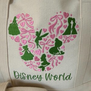 May include: Cream-colored tote bag with a large embroidered design. The design features a Mickey Mouse head shape filled with pink hearts, bows, and green silhouettes of Disney characters. The words "Disney World" are embroidered in green below the design.