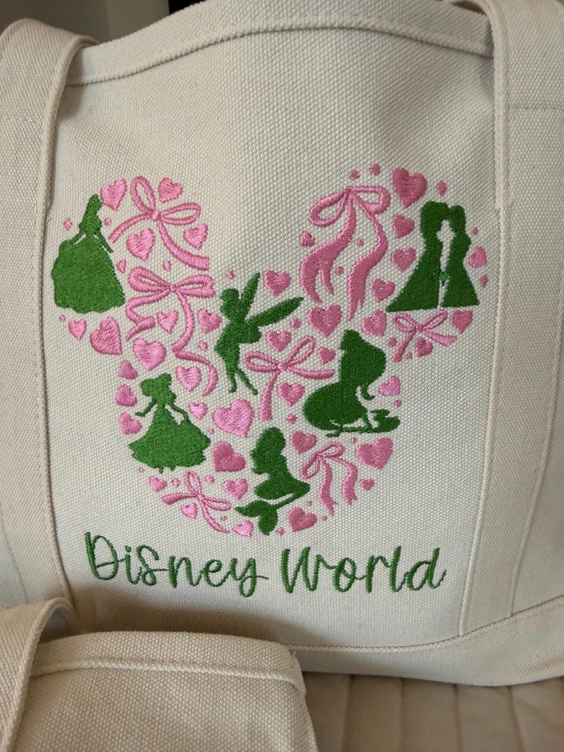May include: Cream-colored tote bag with a large embroidered design. The design features a Mickey Mouse head shape filled with pink hearts, bows, and green silhouettes of Disney characters. The words "Disney World" are embroidered in green below the design.