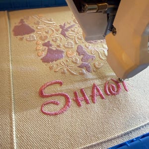 May include: An embroidery machine in operation, stitching a design onto a beige fabric. The design includes pink lettering that spells "SHA@" and decorative elements in pink and light peach. The machine's arm is visible, positioned above the fabric.