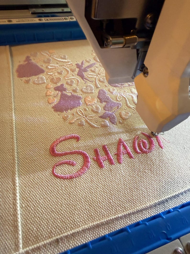 May include: An embroidery machine in operation, stitching a design onto a beige fabric. The design includes pink lettering that spells "SHA@" and decorative elements in pink and light peach. The machine's arm is visible, positioned above the fabric.
