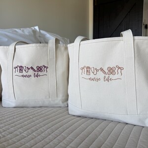 May include: Two off-white canvas tote bags with matching handles. Each bag features a design of medical symbols and the words "nurse life" in a cursive font. The bags are sitting on a bed.
