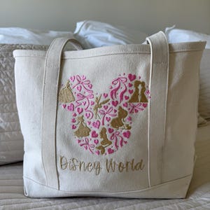 May include: Cream-colored canvas tote bag with embroidered design. The design features a Mickey Mouse head shape filled with pink hearts, bows, and gold silhouettes. The words "Disney World" are embroidered in gold below the design. The bag has two handles.